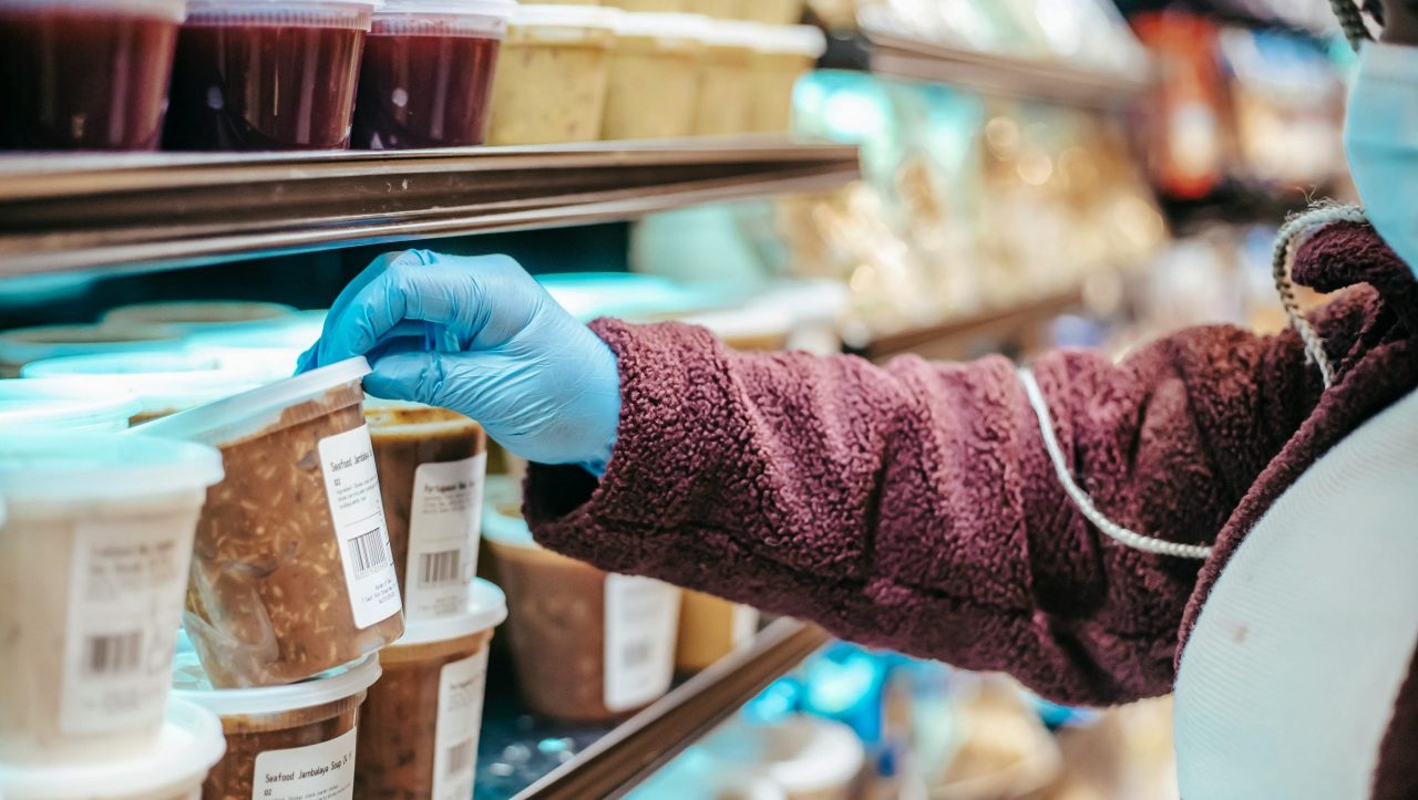 Crop anonymous female customer in protective mask reading label on frozen food in plastic container in grocery store
